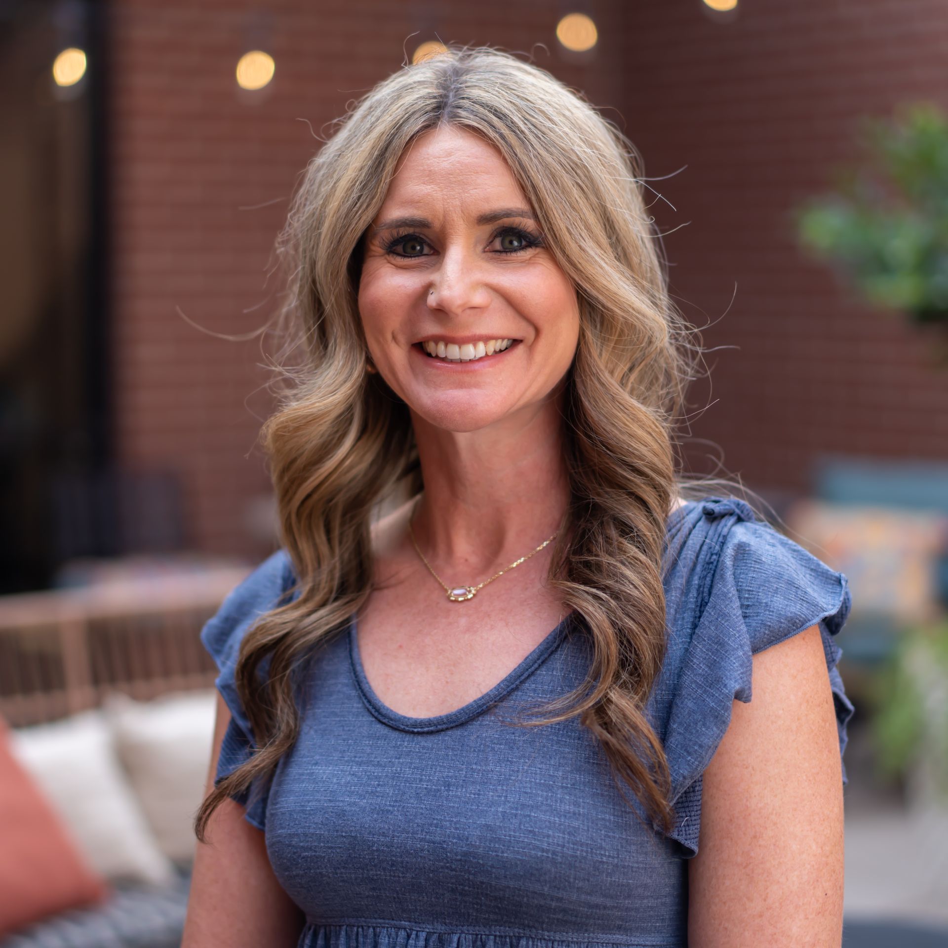 Woman with long, wavy blonde hair smiles outdoors. Wearing a blue top, necklace. Background includes brick and string lights.