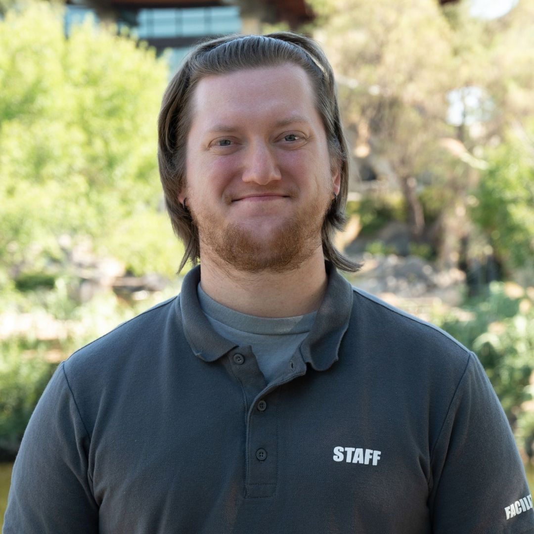 Bald man with glasses and beard, in a blue button-down shirt, smiles outdoors.