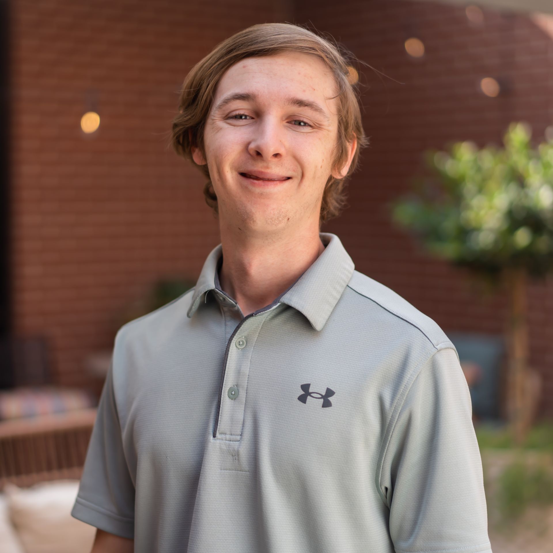 Man with light brown hair, smiling, wearing a gray polo shirt, standing outside with brick wall in the background.