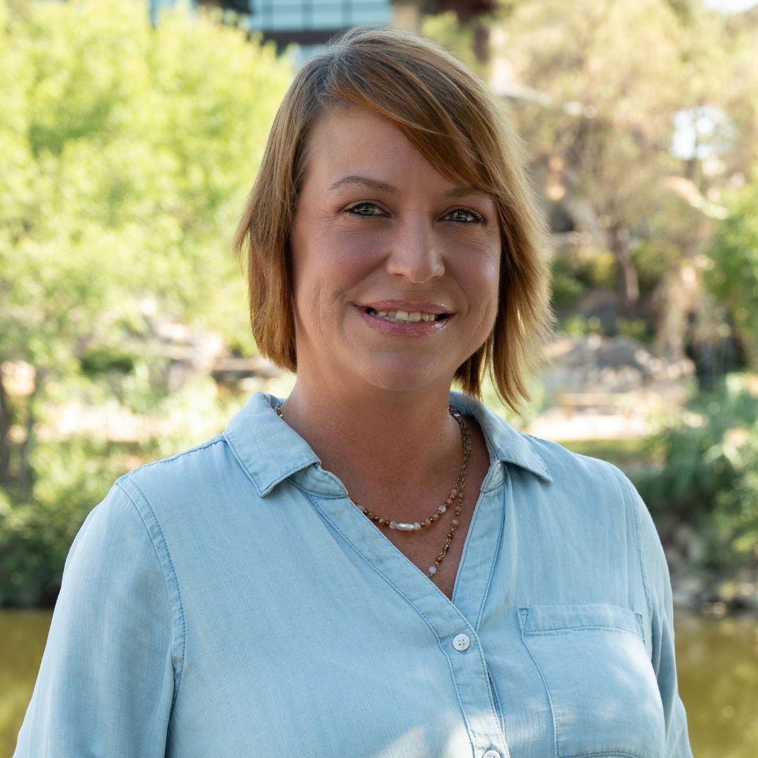Man with a gray beard and blue shirt smiles outdoors. Brick building background with soft lighting.