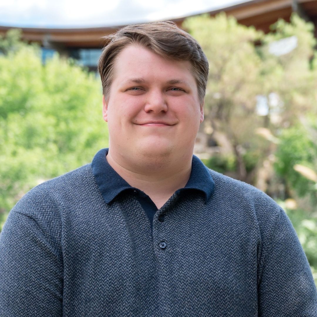 Man with a gray beard and blue shirt smiles outdoors. Brick building background with soft lighting.
