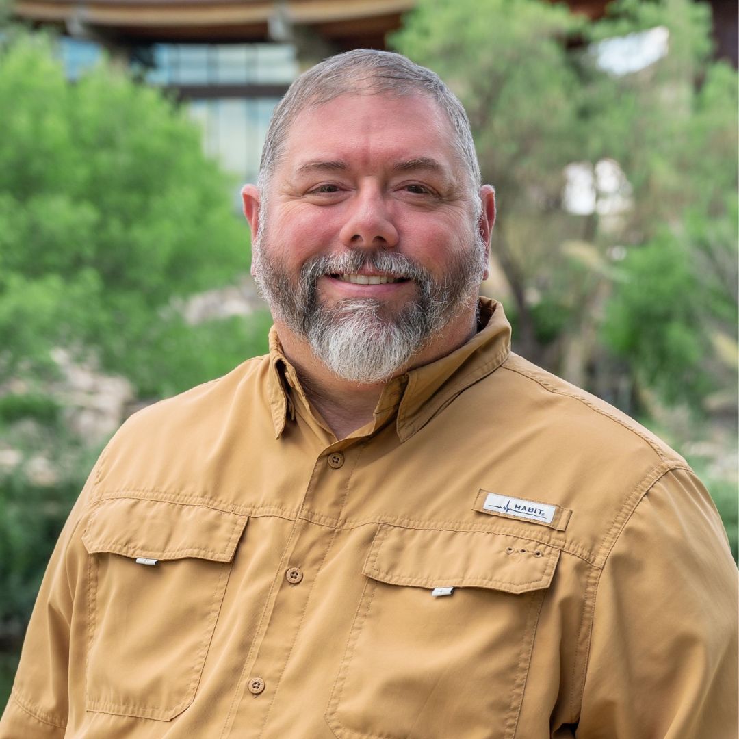 Man with a gray beard and blue shirt smiles outdoors. Brick building background with soft lighting.