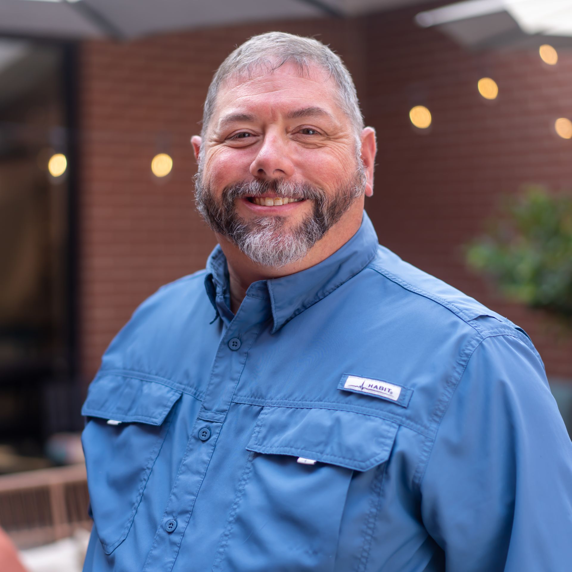 Man with a gray beard and blue shirt smiles outdoors. Brick building background with soft lighting.