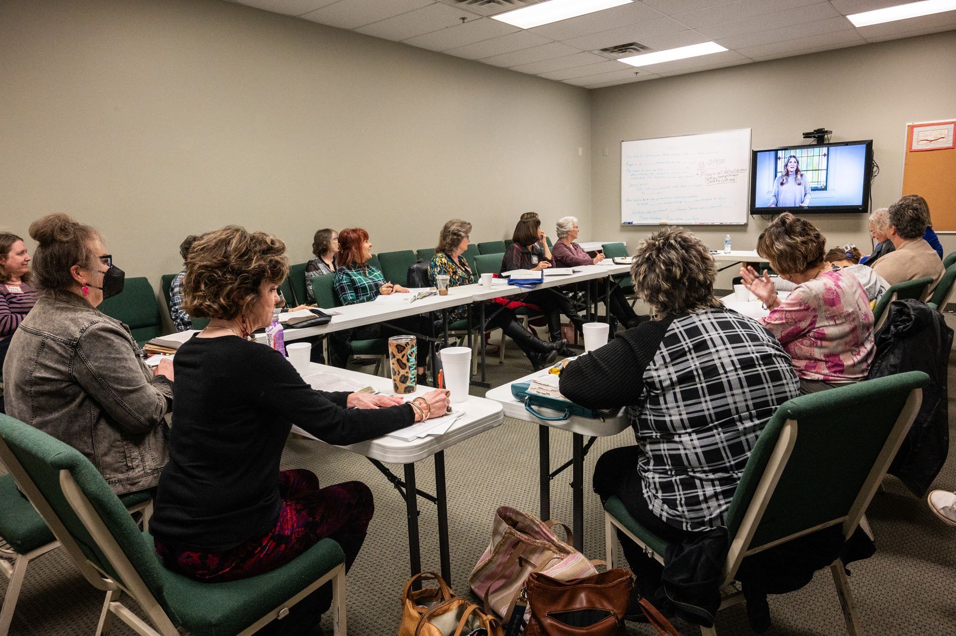 Group of people seated at tables, looking at a whiteboard and a TV screen in a meeting room.