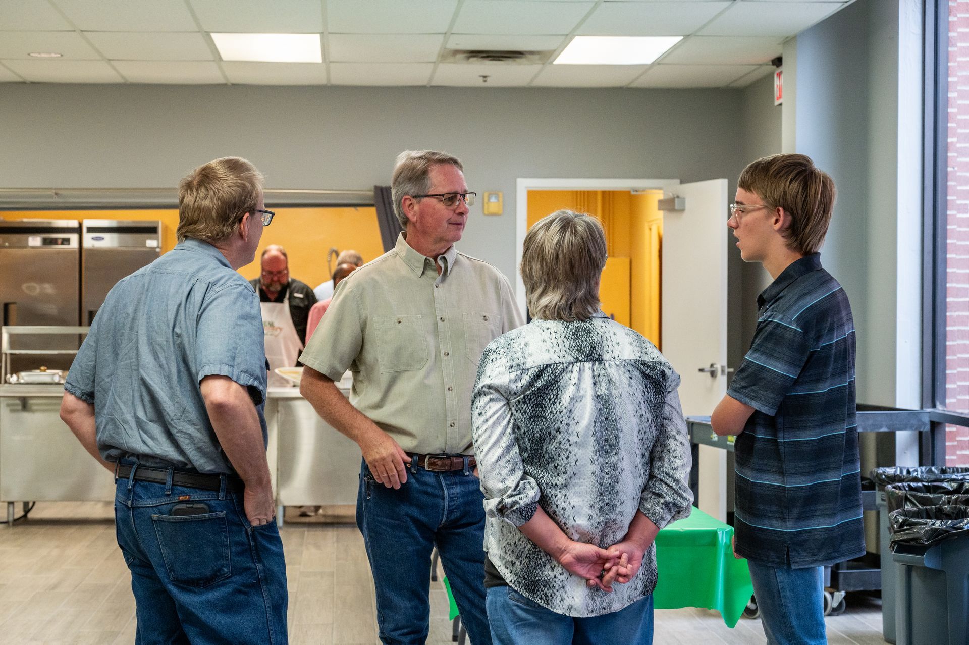 Four people in a room are in conversation. One man wears a light brown shirt, others wear patterned shirts.