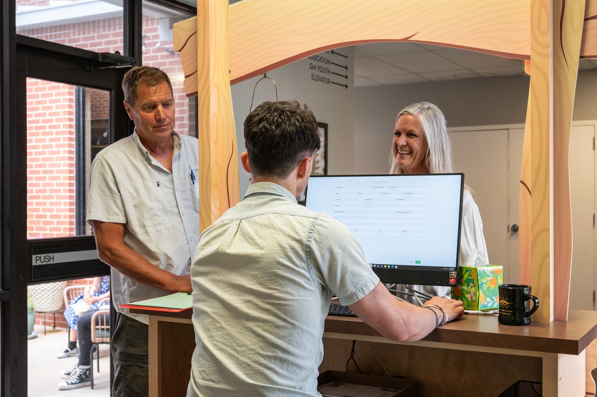 People at a reception desk. A person behind the counter interacts with two others, the monitor displaying text.