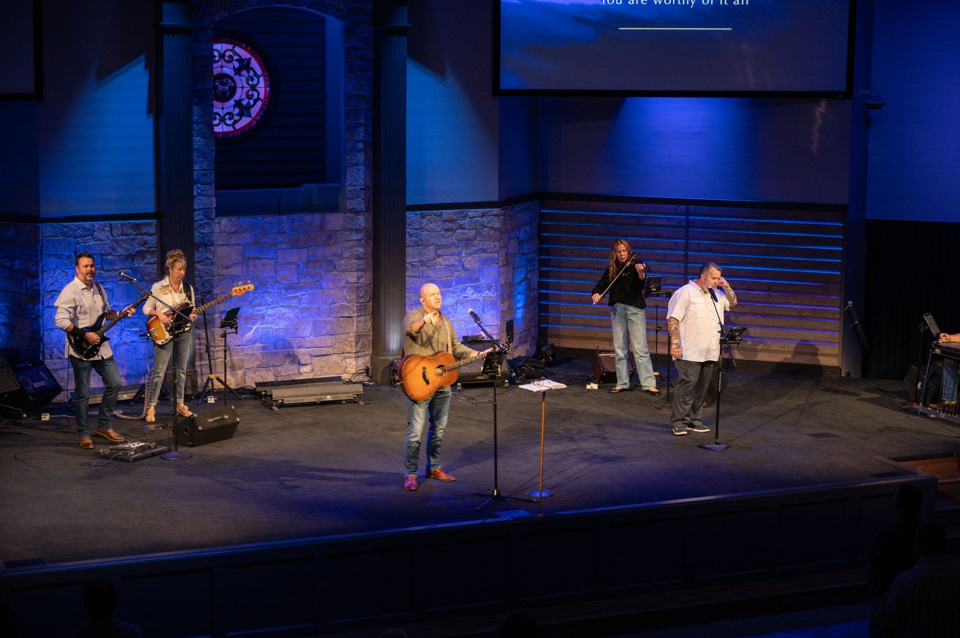 A band performs on a stage in a church. Musicians include a violinist, guitarist, and vocalist. Blue lighting.
