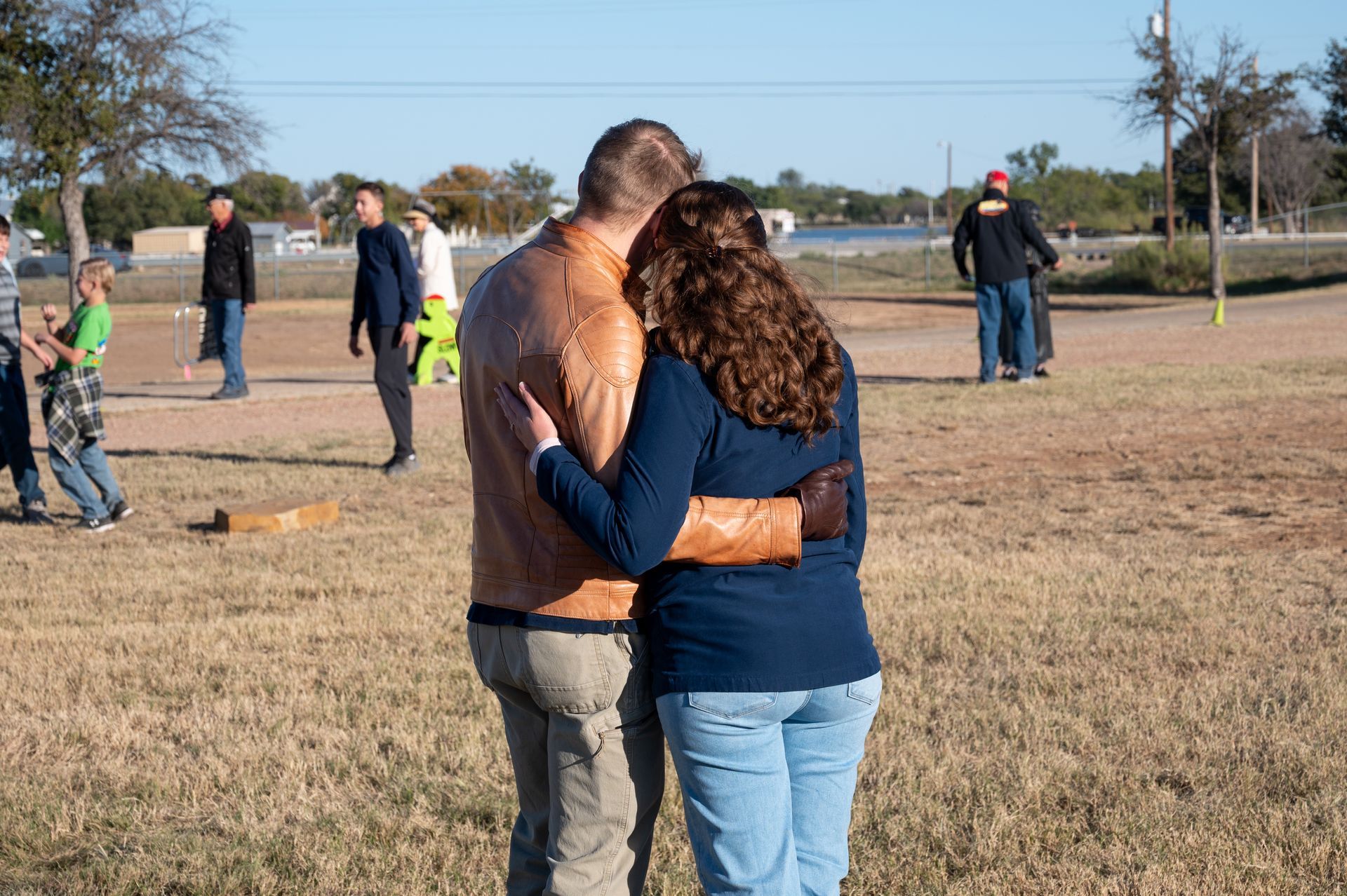 Couple embracing in a grassy field; other people in the background. Sunny day.