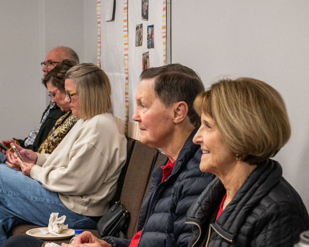 People seated, looking right, in a room with a white wall. Some are smiling, and one holds a phone.