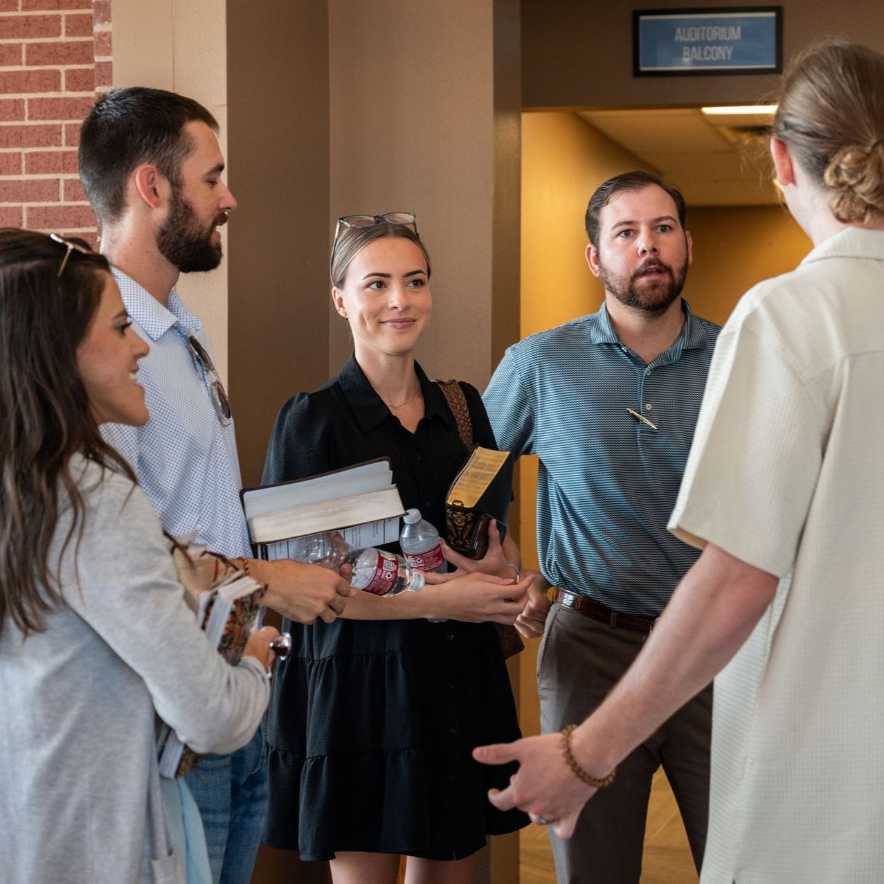 Group of people talking inside a building near a hallway, holding items, possibly on a tour.