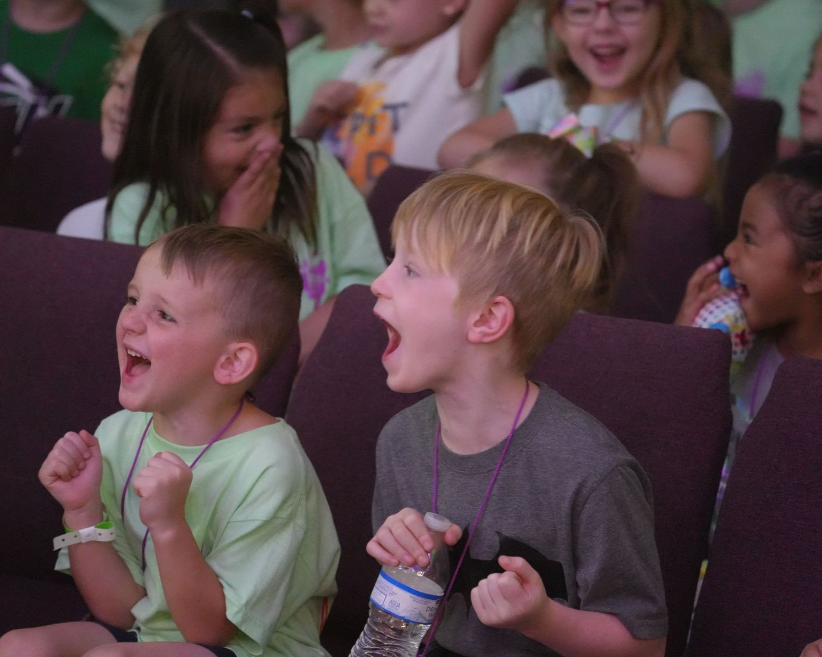 Children cheering, some with open mouths and raised fists, in a room with purple seating.