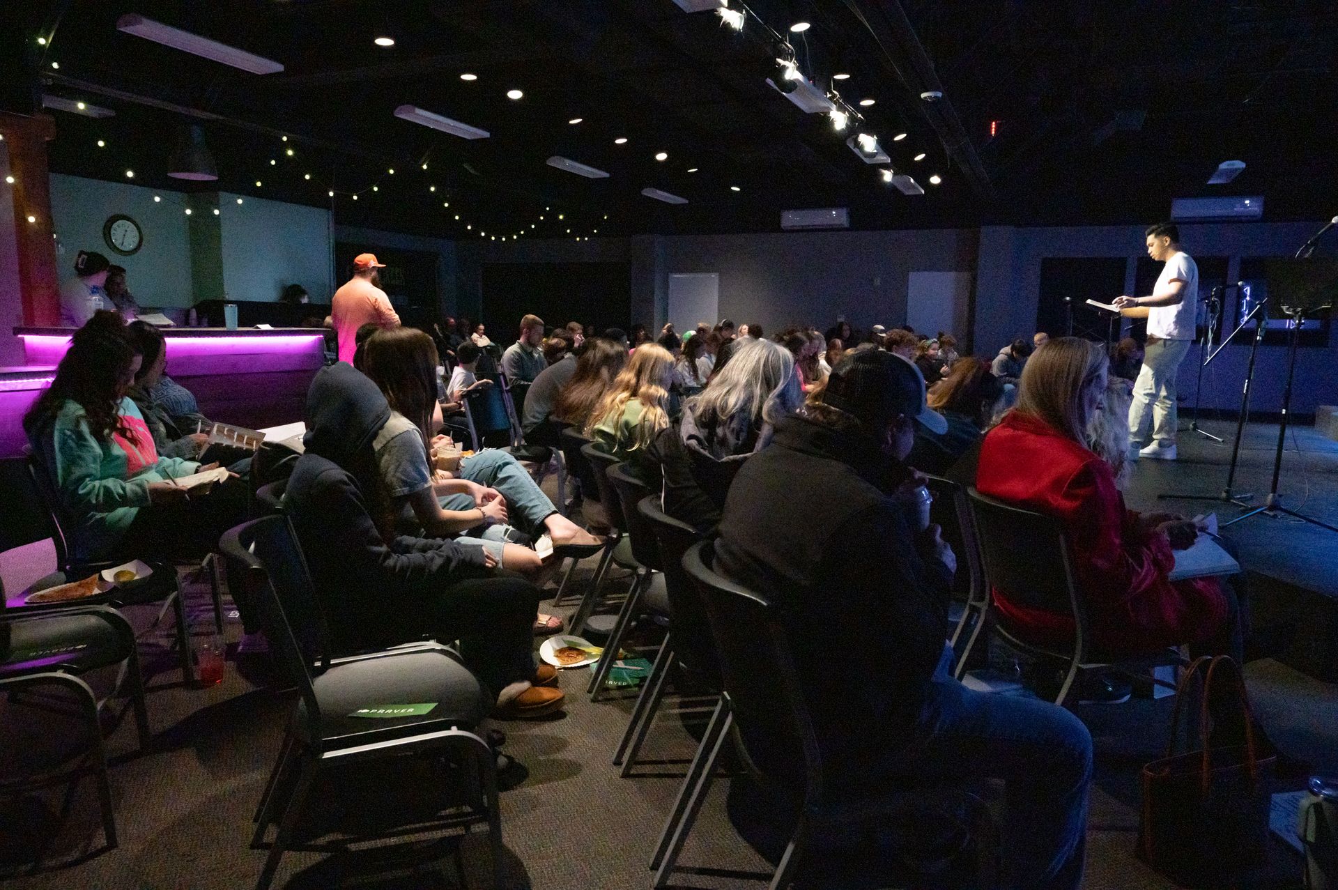 Audience in a dimly lit room, listening to a person on a stage, possibly a talk or performance.