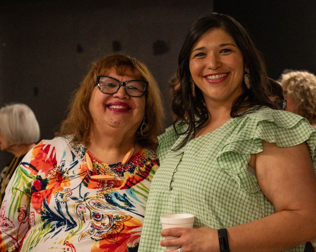 Two smiling people at an event; woman in floral top, woman in green dress holding a cup.