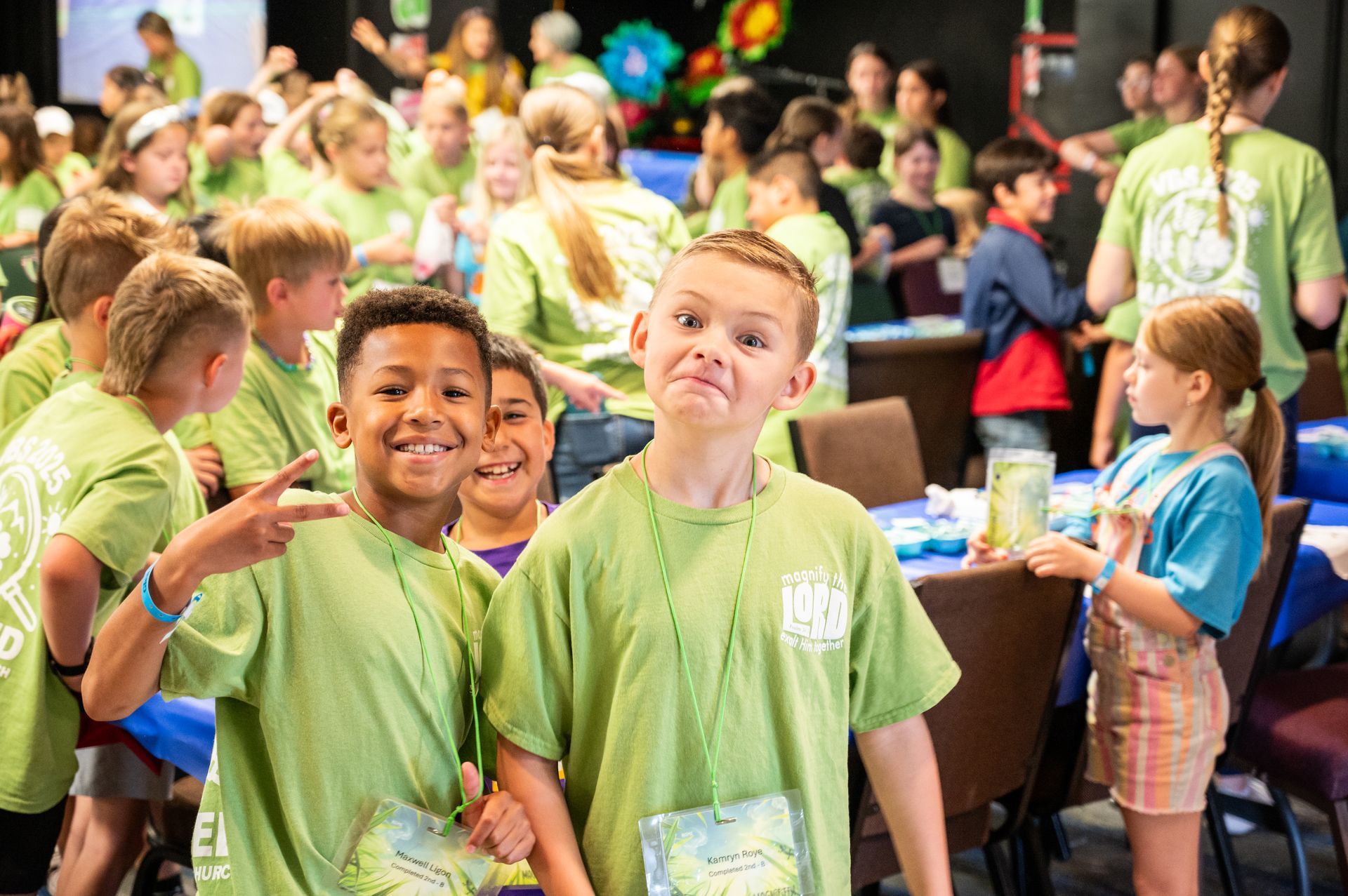 Children and two adults in a classroom with green cabinets, waving and sitting at tables.
