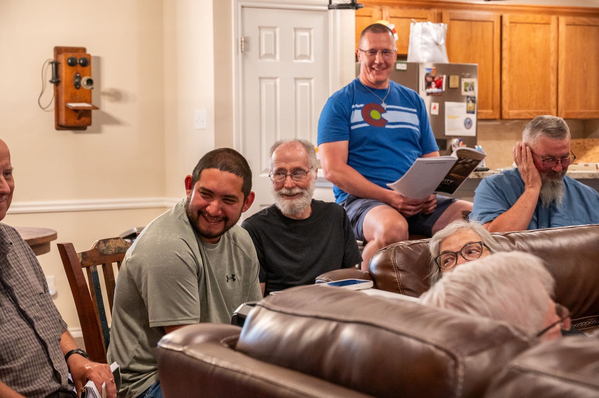 Group of people in a living room, some smiling. Man in blue shirt holds papers, others on couch.