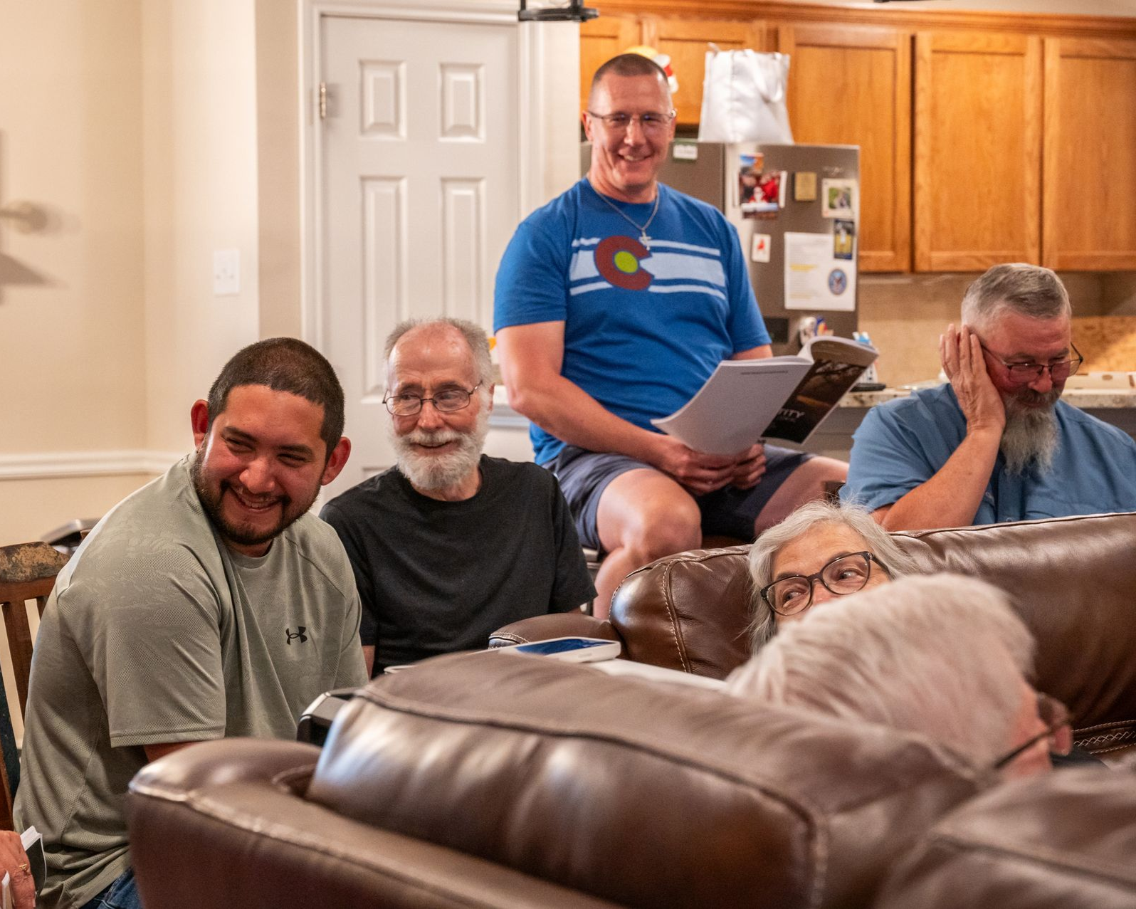 Group of people in a living room; one reading from a paper, others looking on and smiling.
