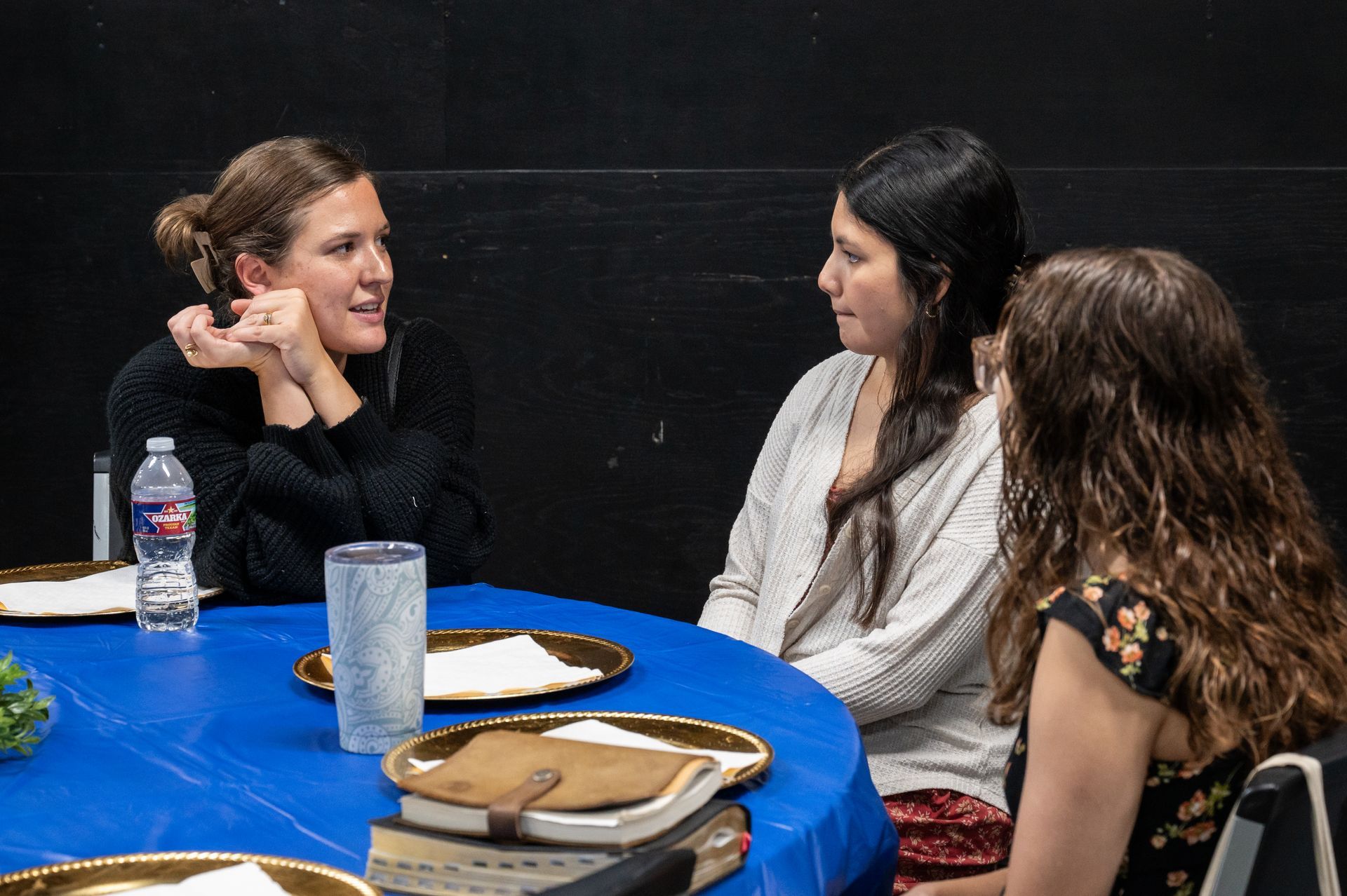 Three people seated at a table, conversing. Blue tablecloth, plates, and a water bottle are visible.
