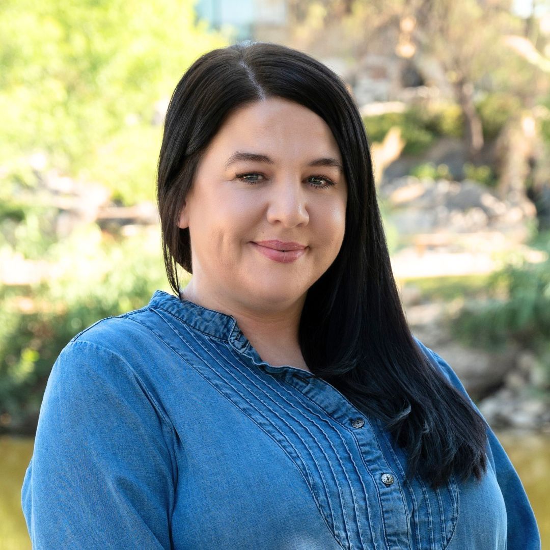 Woman in blue shirt and glasses smiles in front of tall, brown plants.
