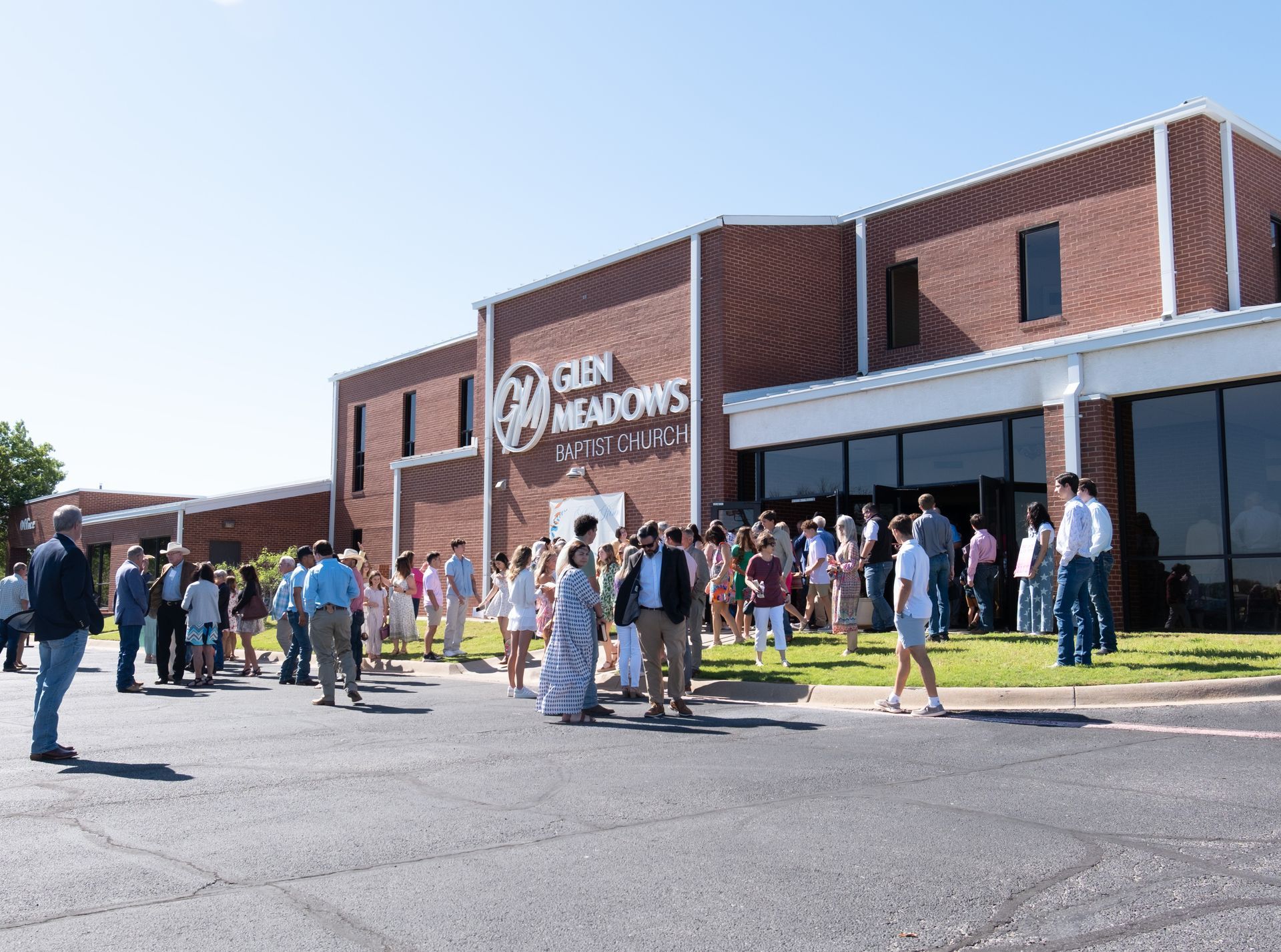 Children and two adults in a classroom with green cabinets, waving and sitting at tables.