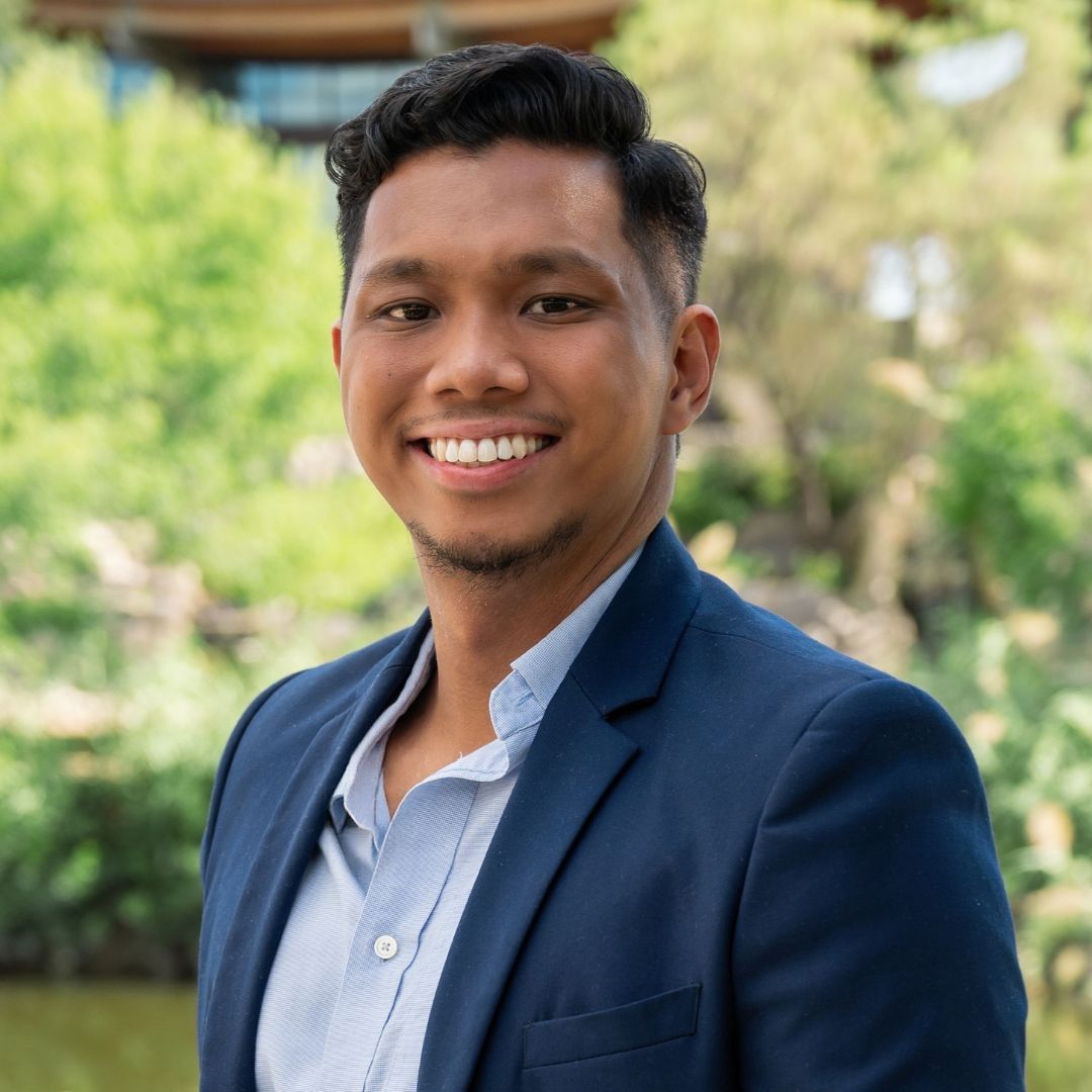 Woman with long, wavy blonde hair smiles outdoors. Wearing a blue top, necklace. Background includes brick and string lights.