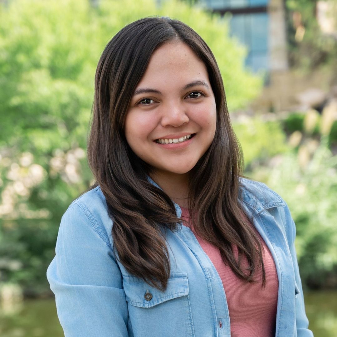 Man with beard, glasses, light blue shirt smiling outdoors with brick wall background.
