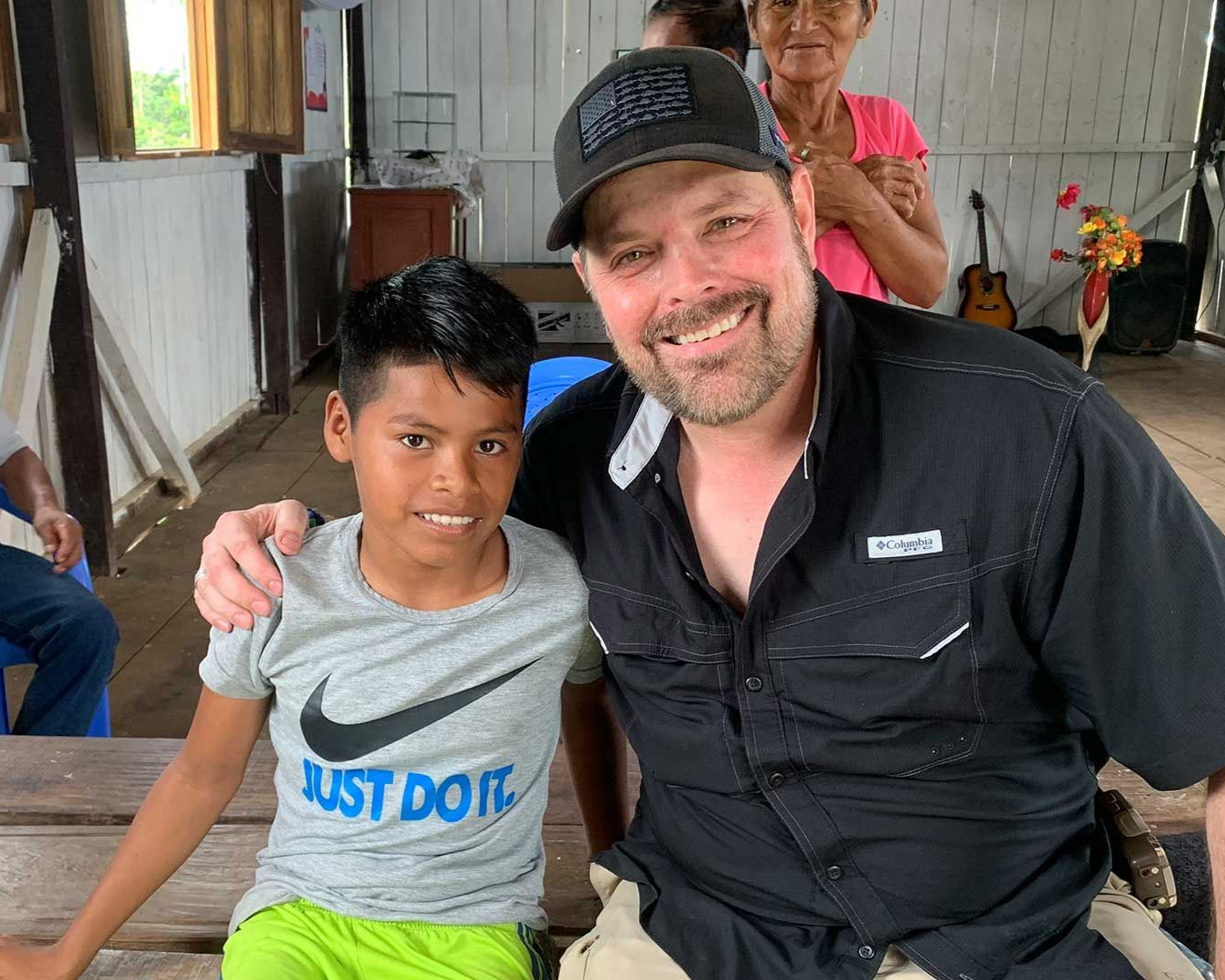 Man with dark cap smiles, arm around boy in gray Nike shirt. Wooden interior; another person in background.