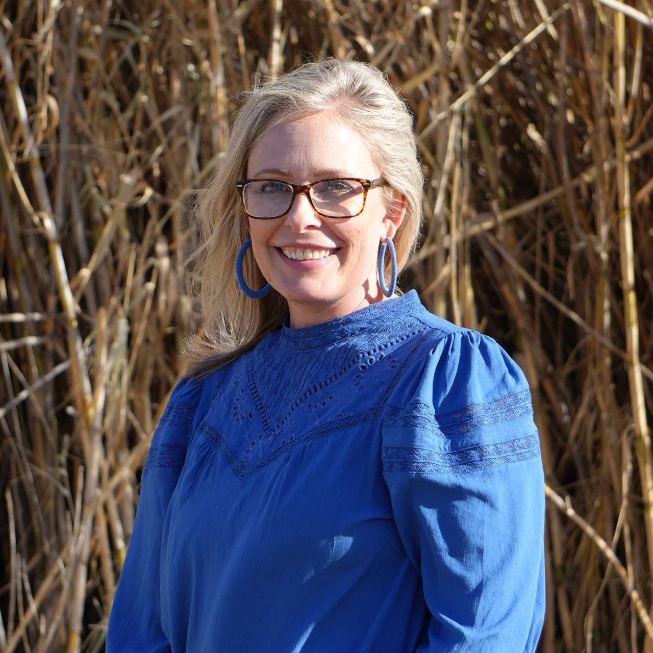 Woman in blue shirt and glasses smiles in front of tall, brown plants.