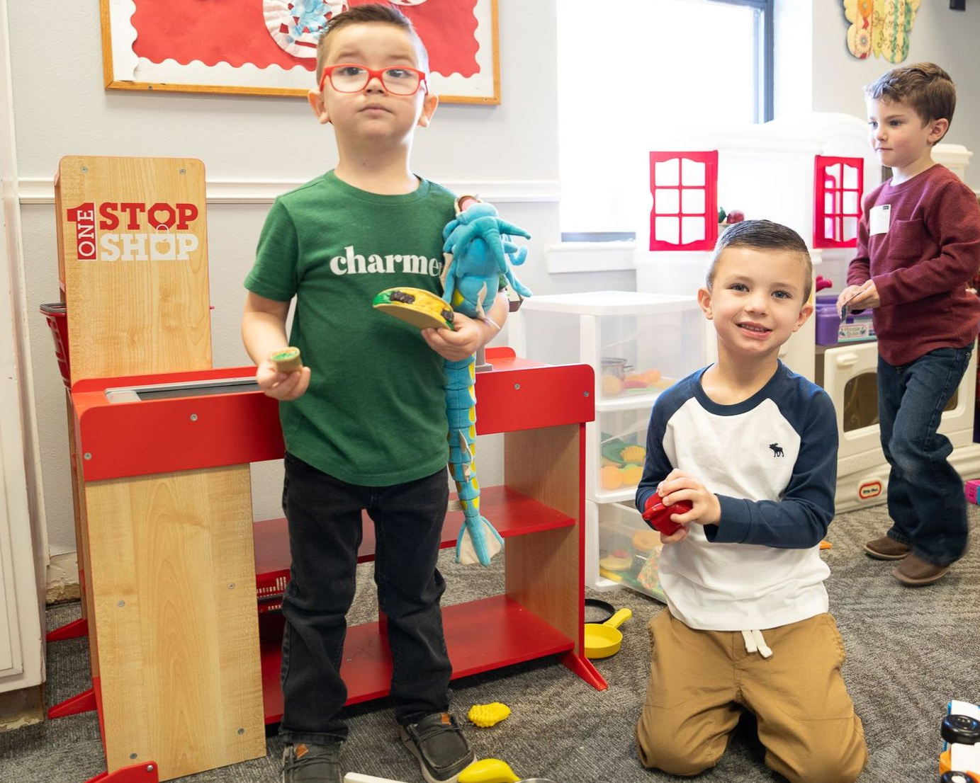 Children playing with a toy shop. One holds a stuffed animal and toy food. Another kneels holding a toy.