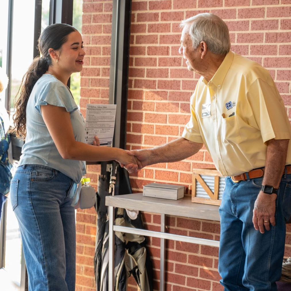 A young person and older person shaking hands in front of a brick wall and open door.