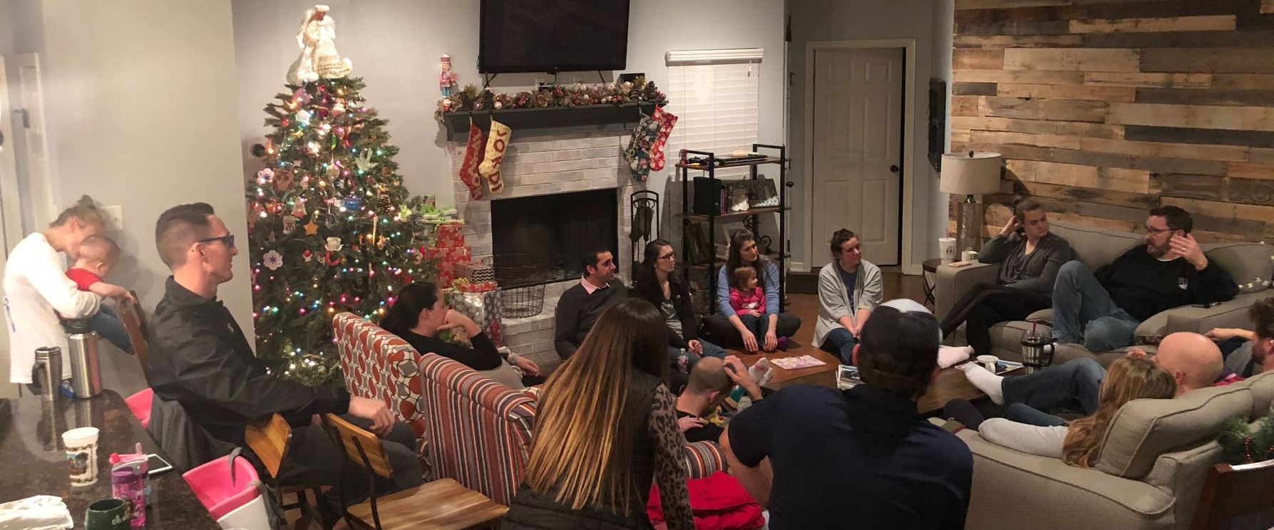 People gather in a living room decorated for Christmas. Some sit on a couch, others on the floor.