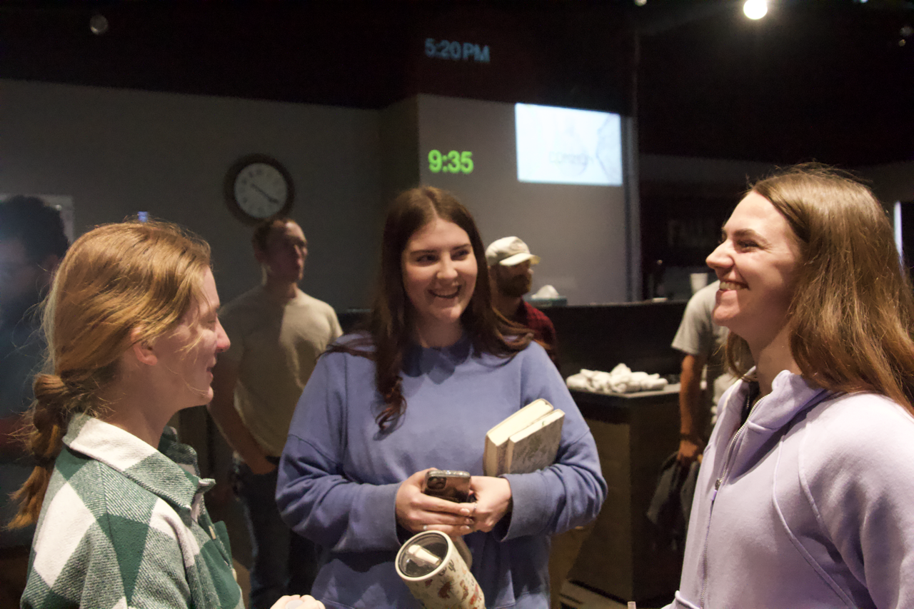 Three people chatting inside; a clock and screen in background.