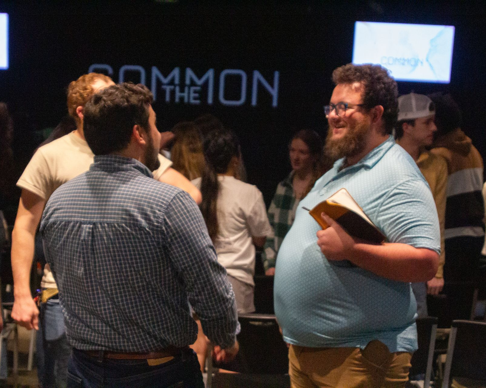 Two men talking in a room with a crowd. One holds a book, smiles.