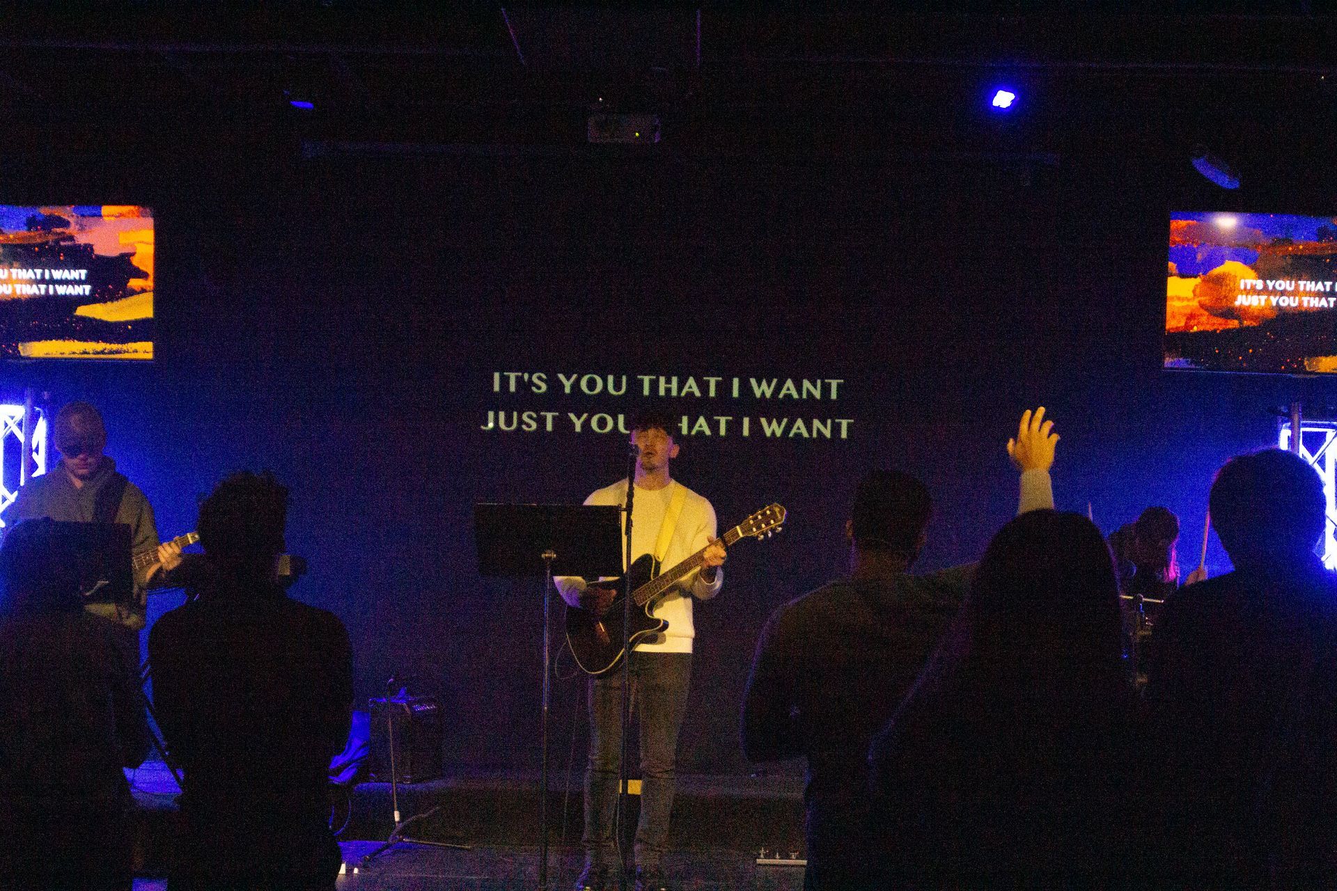 A band performing on stage, lyrics displayed, with people singing and raising hands in worship.