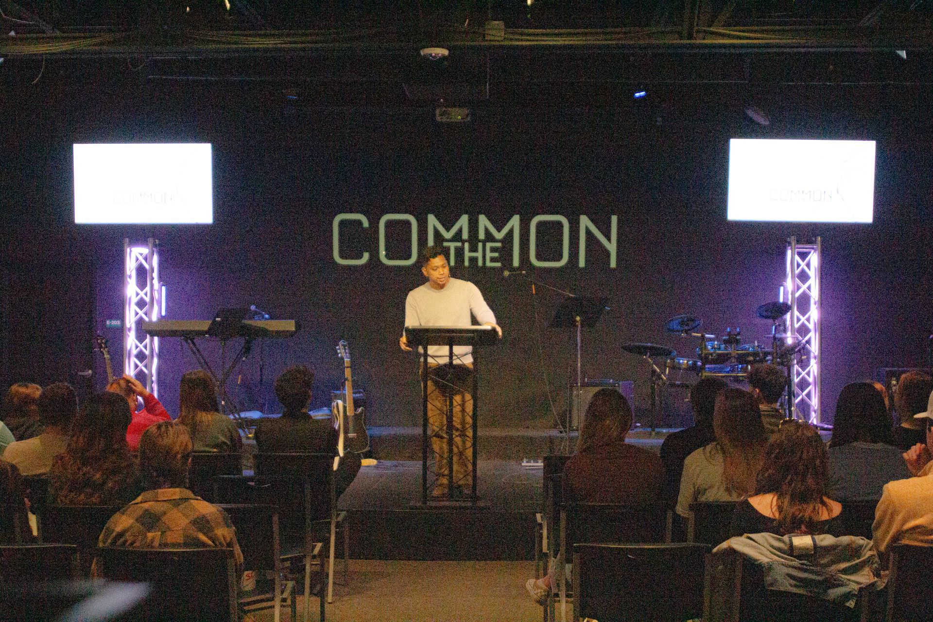 A man speaks at a podium on a stage with the words "Common" displayed. People sit in chairs.