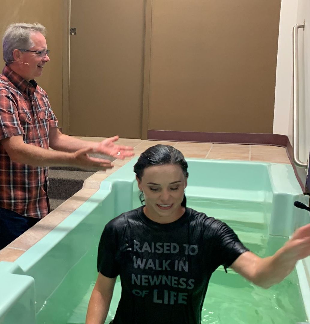 Woman in a baptismal pool, being baptized by a man. The woman is wearing a black shirt, smiling.
