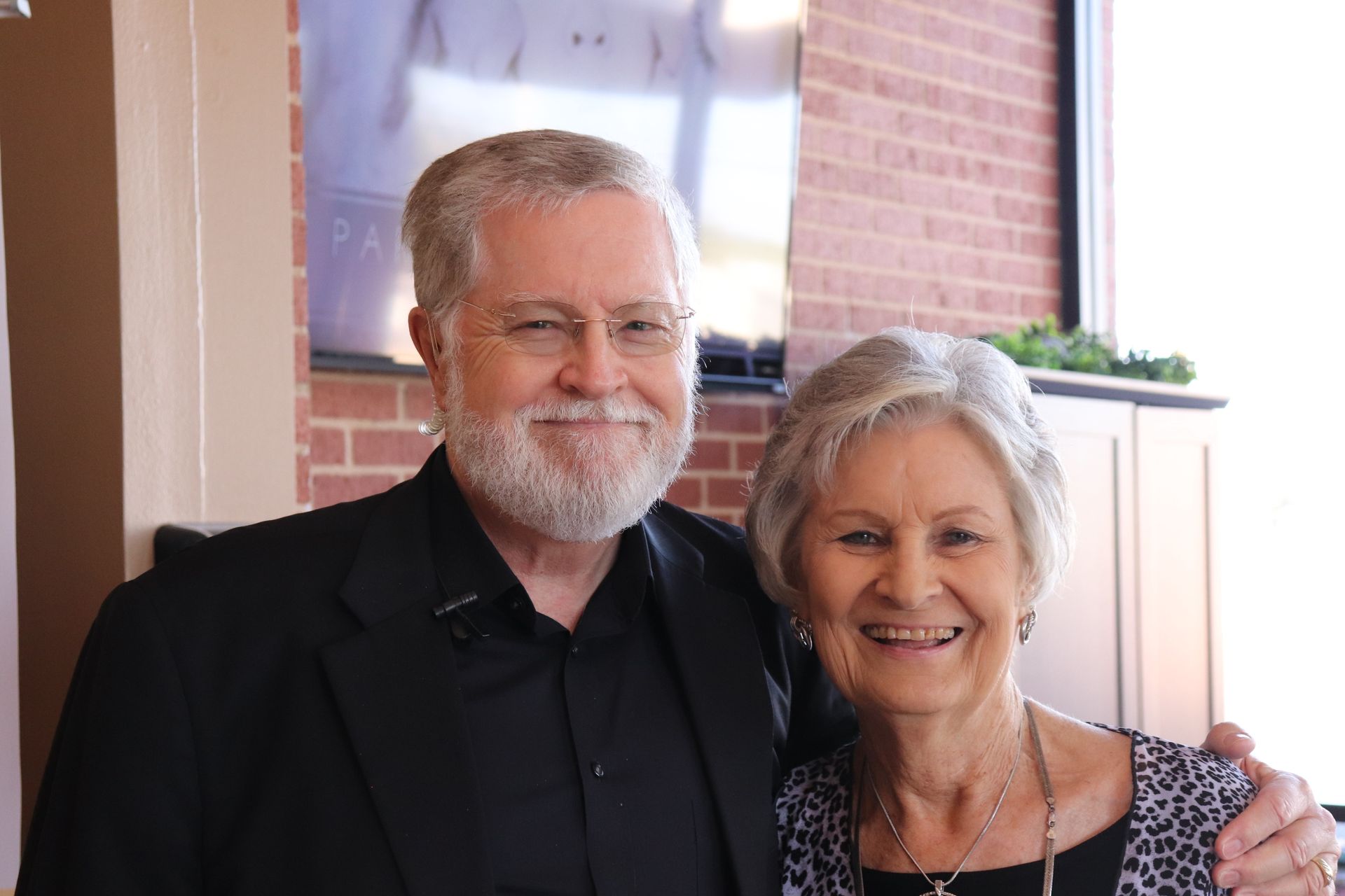 Man with a beard and woman smile, posing together inside a building with a brick wall.