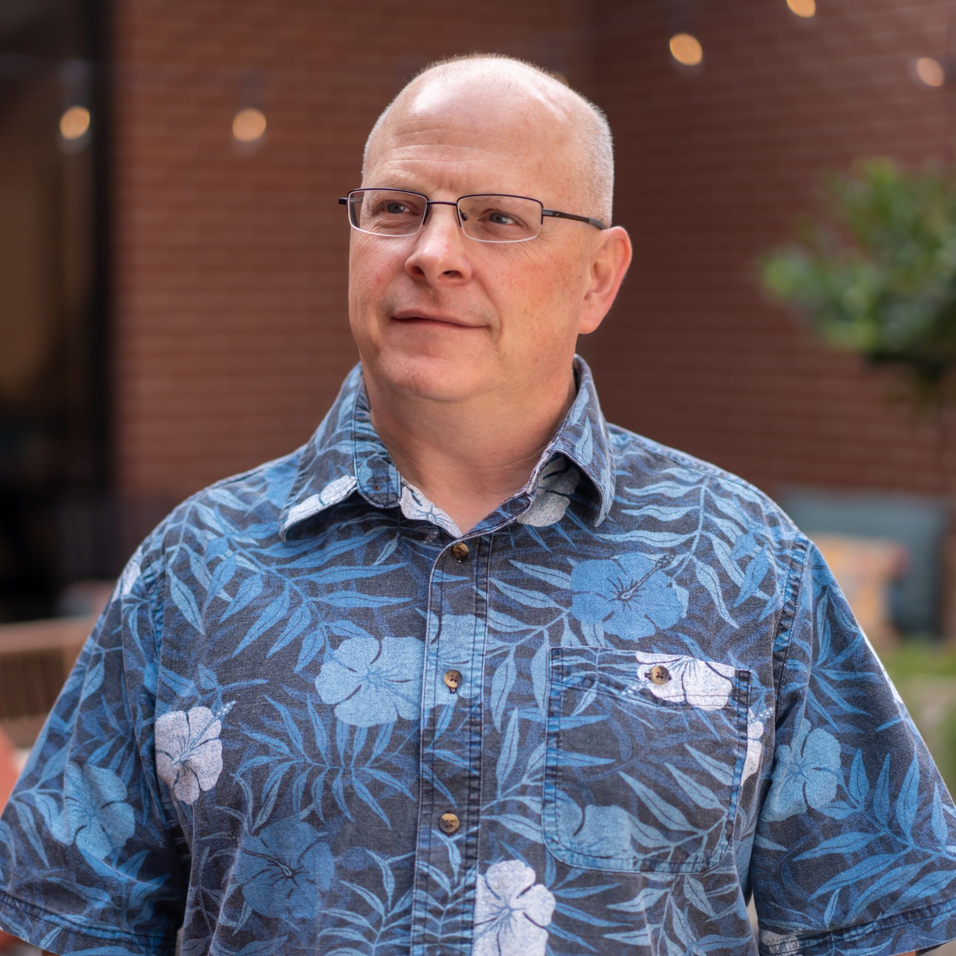 Man with glasses in blue floral shirt, looking off-camera. Outdoors, brick background.
