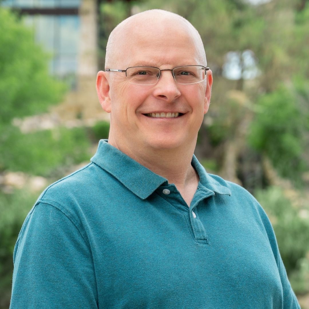 Bald man with glasses and beard, in a blue button-down shirt, smiles outdoors.