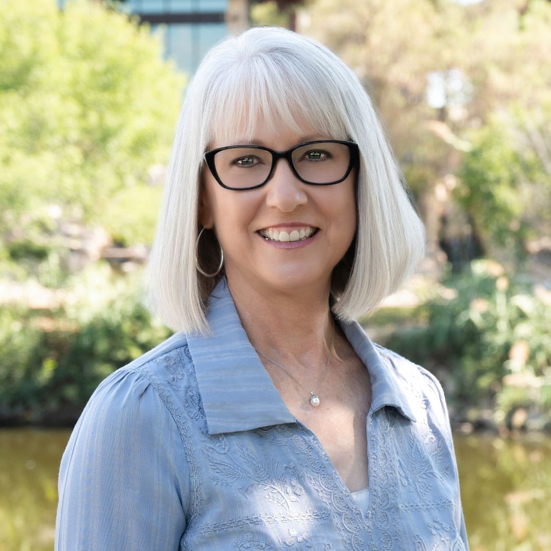 Woman with gray hair and glasses smiling in a courtyard. She wears a navy blue shirt.