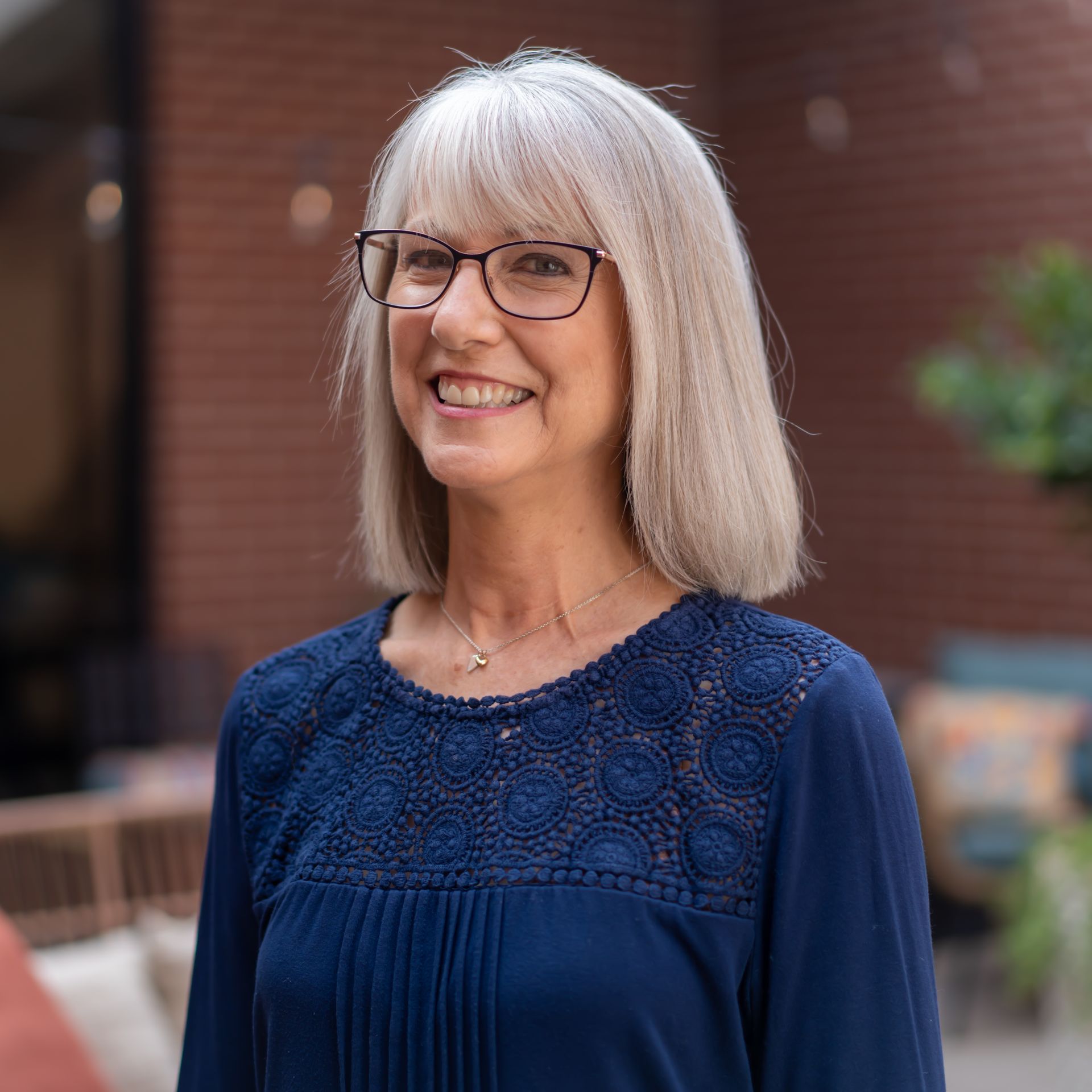 Woman with gray hair and glasses smiling in a courtyard. She wears a navy blue shirt.
