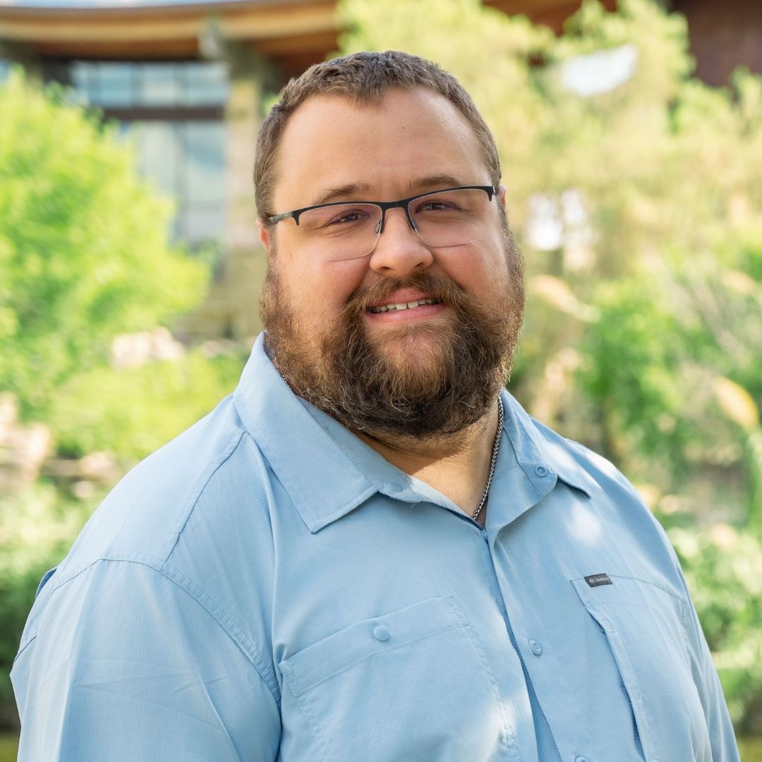 Bald man with glasses wearing a blue shirt with a white cross. Smiling outdoors near brick wall.