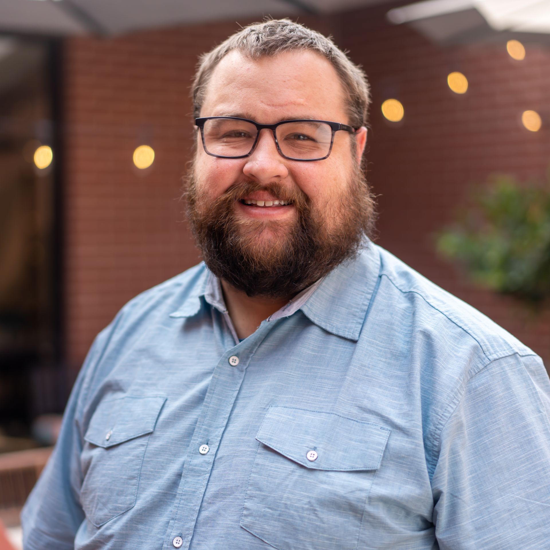 Man with beard, glasses, light blue shirt smiling outdoors with brick wall background.