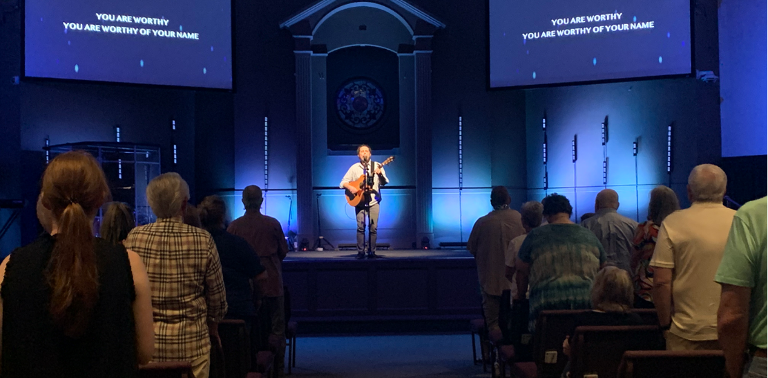 A man playing a guitar on a stage with a church congregation. Blue stage lighting. Lyrics on screens.