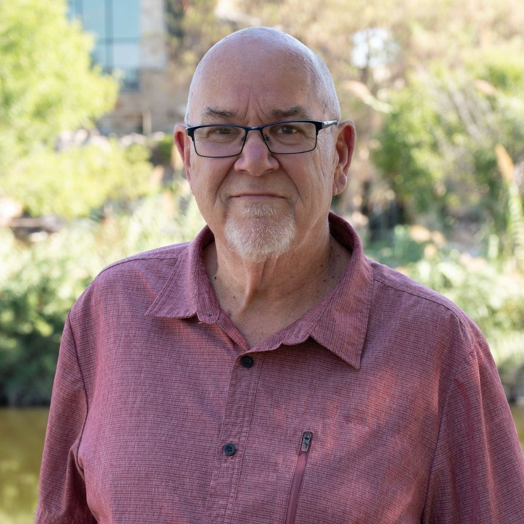 Bald man with glasses wearing a blue shirt with a white cross. Smiling outdoors near brick wall.