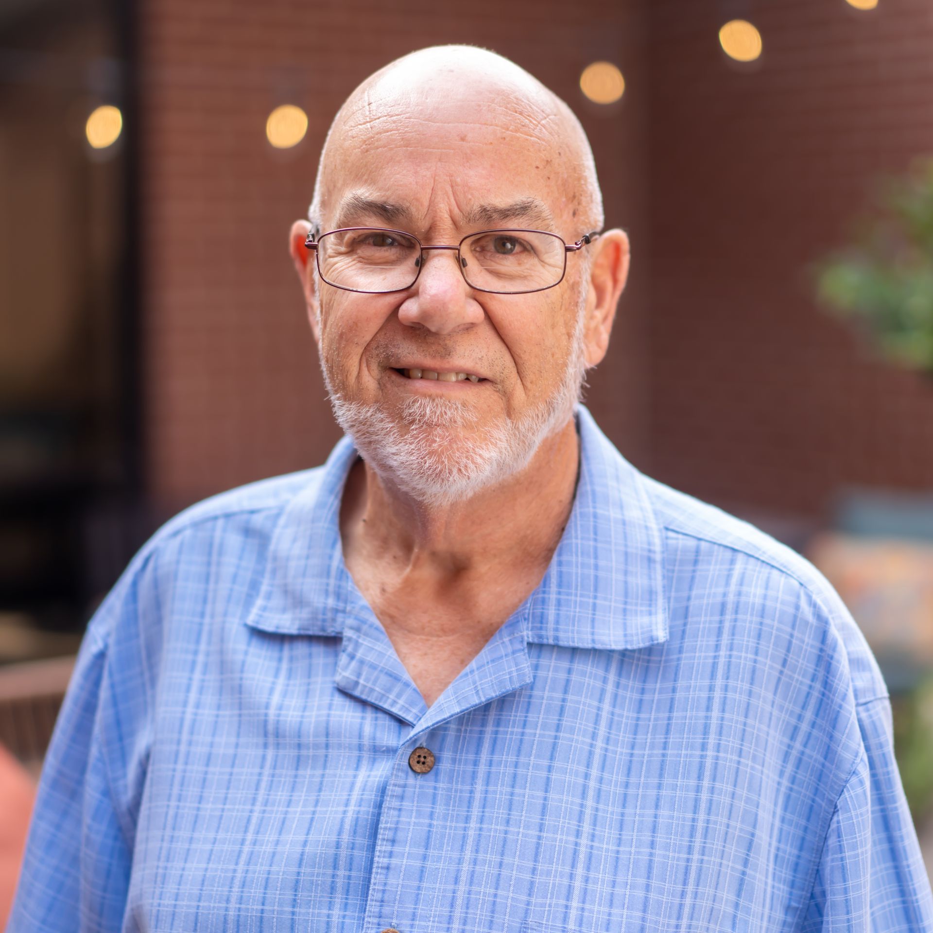 Bald man with glasses and beard, in a blue button-down shirt, smiles outdoors.