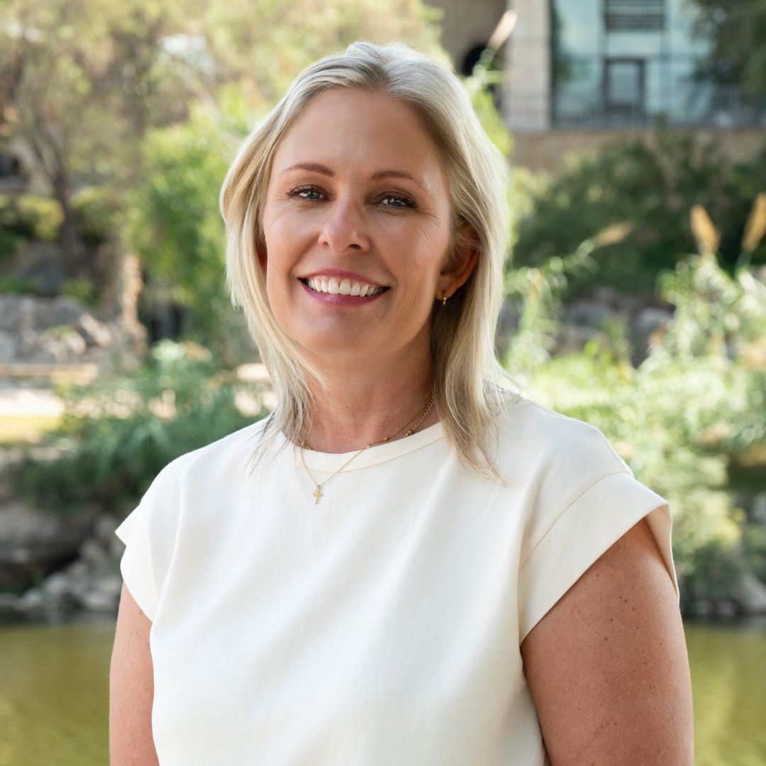 Man with a gray beard and blue shirt smiles outdoors. Brick building background with soft lighting.