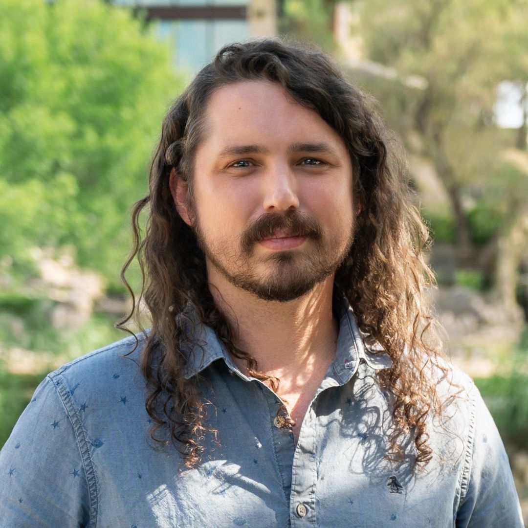 Man with long hair in a bun smiles while wearing a blue patterned shirt outdoors near a brick building.