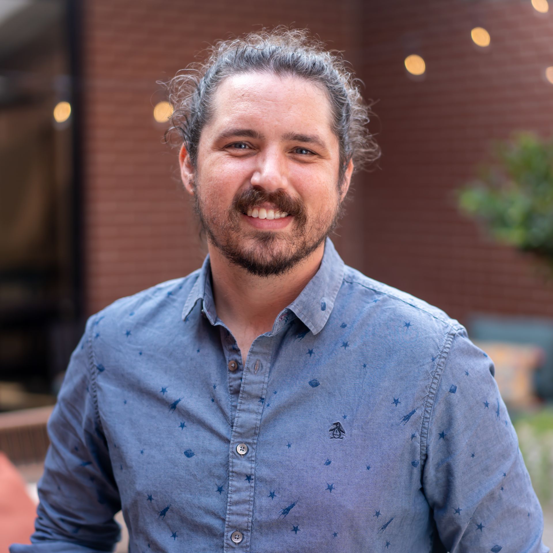 Man with long hair in a bun smiles while wearing a blue patterned shirt outdoors near a brick building.