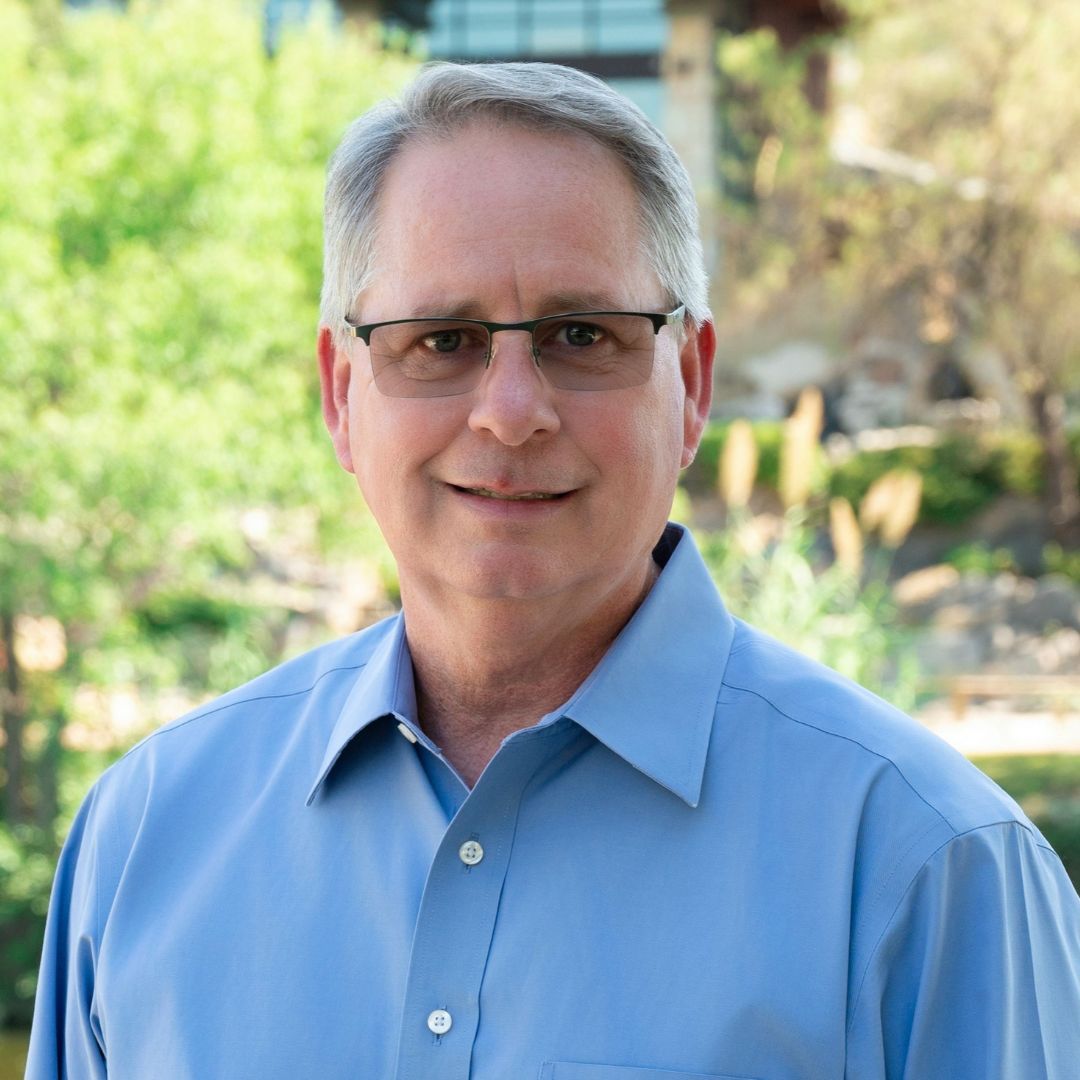 Man in blue patterned shirt and glasses smiles at the camera against a gray backdrop.