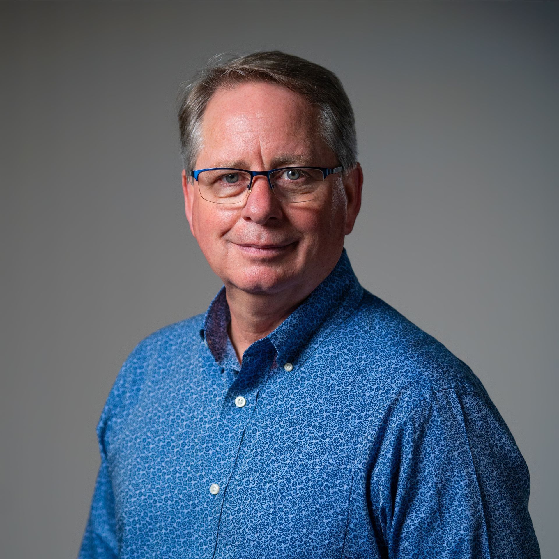Man in blue patterned shirt and glasses smiles at the camera against a gray backdrop.